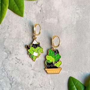 May include: Gold hoop earrings with black cat and plant designs. One earring features a cat peeking out of a geometric planter, the other a cat in a plant pot. The earrings have green and white accents.