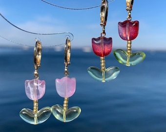 Puede incluir: Dos pares de pendientes de aro dorados con colgantes en forma de tulipán. Un par tiene flores de color rosa translúcido con hojas verde claro, mientras que el otro tiene flores de color rojo intenso con hojas verde claro, con pequeñas cuentas doradas.