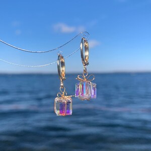 Könnte beinhalten: Ein Paar goldene Creolen mit Anhänger in Form von Geschenkboxen aus schimmerndem Kristall. Die Ohrringe sind vor einem Hintergrund aus blauem Himmel und Wasser fotografiert.