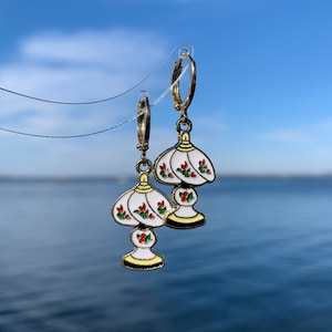 May include: A pair of gold hoop earrings with white lampshade charms. The lampshades have red and green floral details.