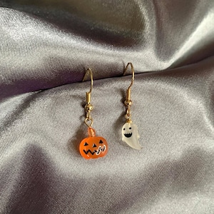 以下が含まれることがあります： ハロウィンをテーマにした金色のイヤリングのペア。片方のイヤリングは、黒いジャック・オー・ランタンの顔をした小さなオレンジ色のカボチャ。もう片方のイヤリングは、黒い笑顔の顔をした白い幽霊です。