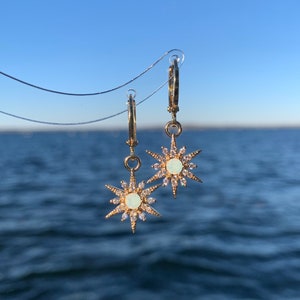 May include: Gold hoop earrings with a dangling star charm. The star is adorned with clear rhinestones and a white opal in the center.