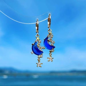May include: Gold hoop earrings with a blue crescent moon charm and a dangling star charm. The charms are decorated with small clear rhinestones.