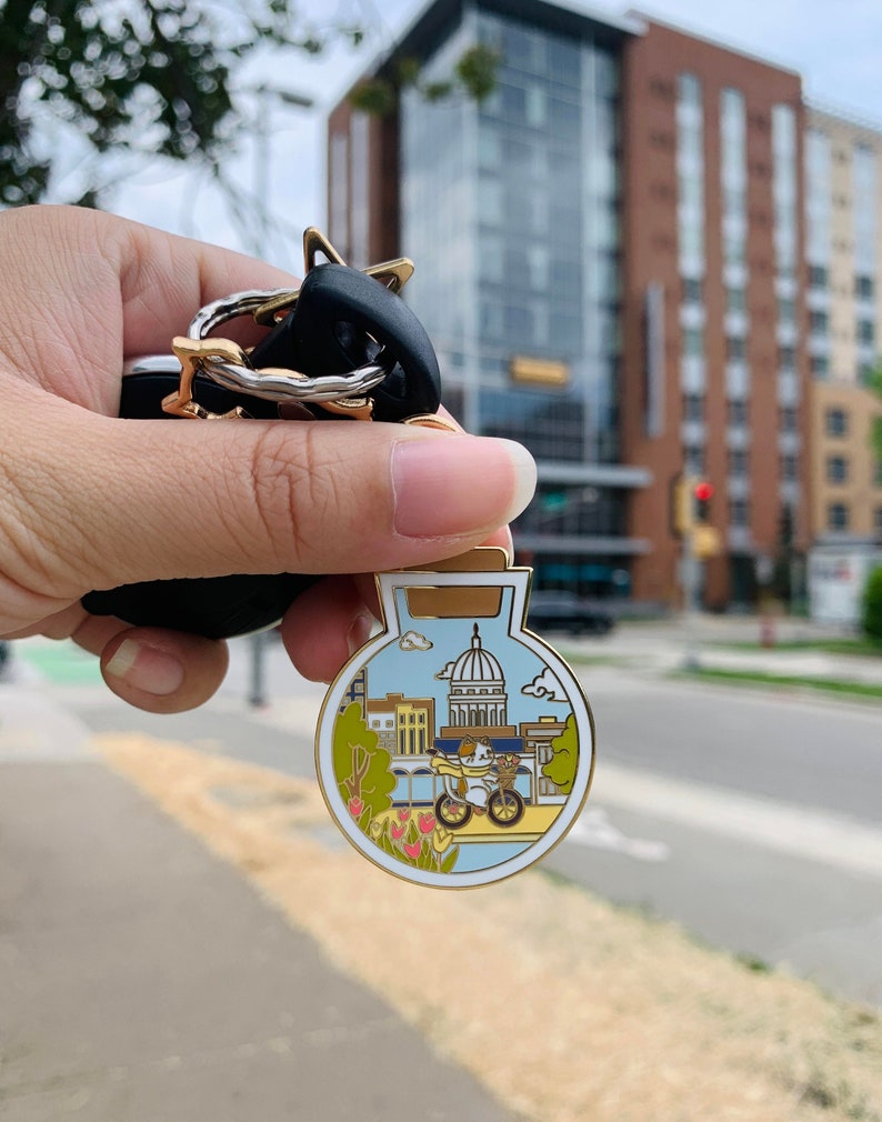 Pu&ograve; includere: Uno spillo in smalto color oro a forma di bottiglia di vetro con un paesaggio urbano in blu e bianco all'interno. Il paesaggio urbano presenta un edificio con una cupola, una bicicletta e una persona che va in bicicletta. Lo spillo &egrave; circondato da un bordo bianco e ha un bordo dorato.