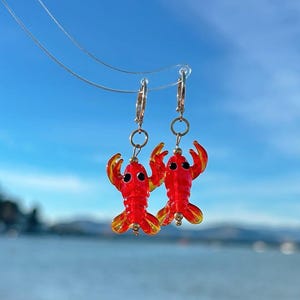 Può includere: Un paio di orecchini a forma di aragosta. Gli orecchini sono rossi con dettagli gialli sulle chele e sulla coda. Sono appesi a cerchi dorati. Lo sfondo è un cielo blu brillante e acqua.