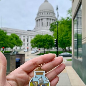 Pu&ograve; includere: Un ciondolo in metallo dorato a forma di bottiglia con sfondo bianco. La bottiglia contiene una rappresentazione in miniatura del Campidoglio dello Stato del Wisconsin, un gatto che guida una bicicletta e un motivo floreale.