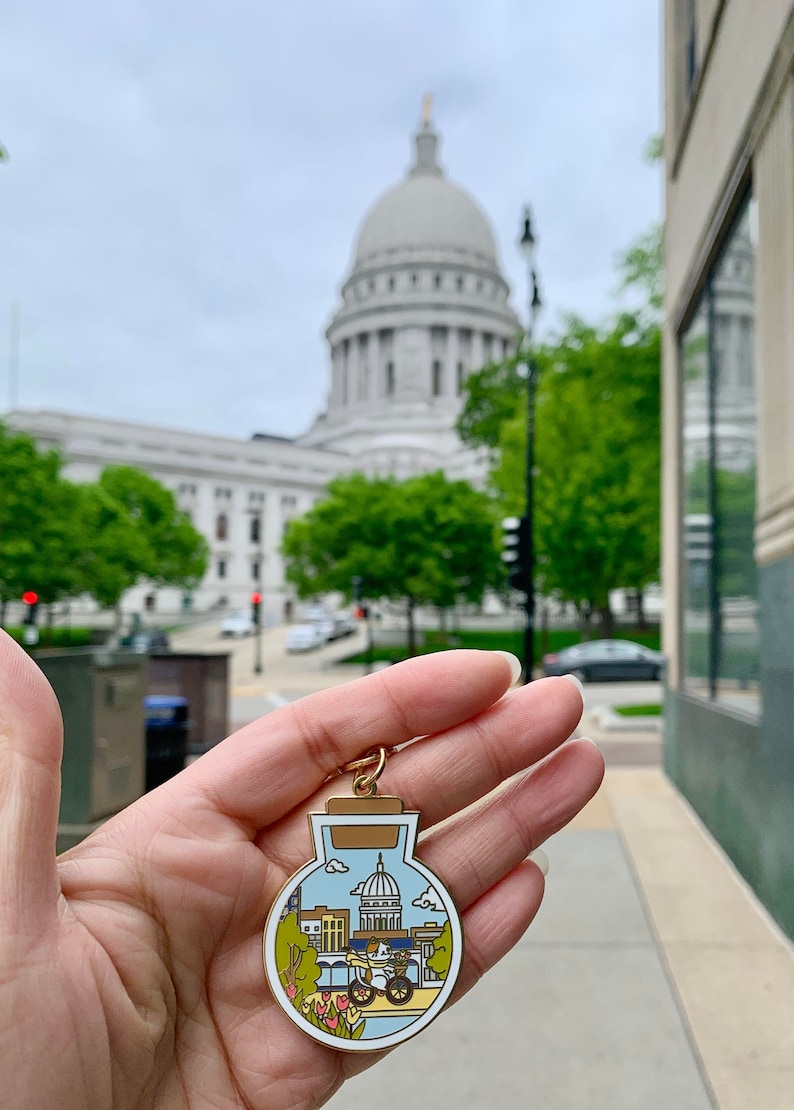 Pu&ograve; includere: Un ciondolo in metallo dorato a forma di bottiglia con sfondo bianco. La bottiglia contiene una rappresentazione in miniatura del Campidoglio dello Stato del Wisconsin, un gatto che guida una bicicletta e un motivo floreale.