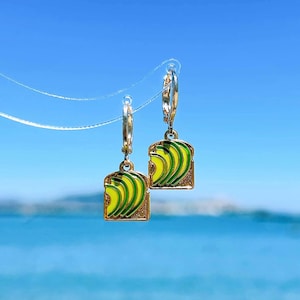 May include: A pair of gold-colored hoop earrings with avocado toast charms. Each charm features a slice of toast with green and yellow avocado slices, outlined in dark green. The background is a bright blue sky.