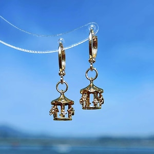 May include: A pair of gold hoop earrings with a small gold carousel charm hanging from each hoop.