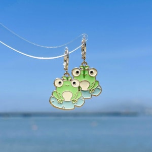 May include: A pair of gold-colored hoop earrings featuring a green frog design. Each frog has large, round eyes and sits on a light blue lily pad. The earrings are set against a blue sky background.