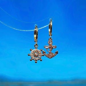 Peut inclure: Une paire de boucles d'oreilles en or sur le thème nautique. Une boucle d'oreille présente une petite ancre avec des strass clairs, tandis que l'autre boucle d'oreille a un petit volant de bateau avec des strass clairs.