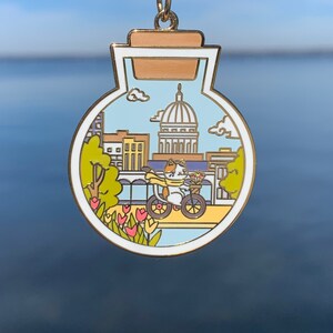 Pu&ograve; includere: Uno spillo in smalto color oro a forma di barattolo di vetro con tappo di sughero. All'interno del barattolo c'&egrave; un'illustrazione di cartone animato di uno skyline urbano con un gatto che guida una bicicletta. Il gatto indossa una sciarpa e porta un cestino di fiori. Lo skyline urbano include un grande edificio con una cupola.