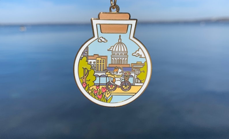 Pu&ograve; includere: Uno spillo in smalto color oro a forma di barattolo di vetro con tappo di sughero. All'interno del barattolo c'&egrave; un'illustrazione di cartone animato di uno skyline urbano con un gatto che guida una bicicletta. Il gatto indossa una sciarpa e porta un cestino di fiori. Lo skyline urbano include un grande edificio con una cupola.