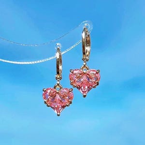 Puede incluir: Un par de pendientes de aro dorados con colgantes de cristal rosa en forma de corazón.