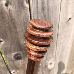 May include: A dark brown wooden honey dipper featuring a series of five graduated circular ridges at the top. The wood has a smooth, polished finish with visible grain patterns throughout the handle and the tiered head of the utensil.
