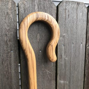 May include: A wooden shepherd's crook featuring a smooth, polished finish with visible natural grain patterns. The top of the staff curves into a rounded hook shape, showcasing the light and dark brown tones of the wood grain throughout the surface.