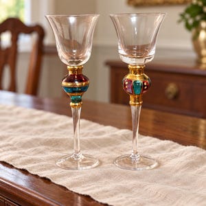 May include: Two clear glass goblets featuring tall, slender stems with decorative, bulbous centers. The stems are accented with gold, deep red, and teal enamel detailing. Each goblet has a wide, flared bowl and a circular base for stability.