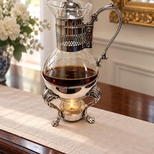 May include: A silver-plated glass coffee carafe with a bulbous body, a decorative filigree collar, and a curved handle. The carafe rests on a silver-plated stand with three ornate legs and a central circular holder containing a lit tea light candle.