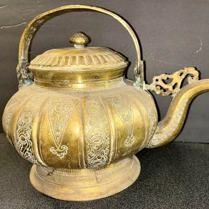 May include: Antique brass teapot with intricate engraved floral patterns. The teapot features a curved spout, a decorative handle, and a lid with a small knob. The teapot sits on a round base and has a tarnished, aged appearance.