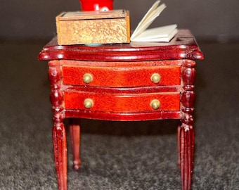 May include: A miniature wooden side table with two drawers and turned legs. The table is painted a rich red color and has gold-colored drawer pulls. A small red box with a lid and a stack of papers are on top of the table.