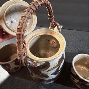May include: Ceramic teapot and two matching cups. The teapot has a woven handle and a lid. The teapot and cups are decorated with brown and blue abstract designs. The interior of the teapot has a built-in strainer.