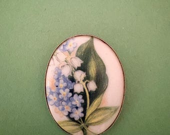 oval brooch featuring a painted floral design of blue forget-me-nots and white lily-of-the-valley flowers with green leaves