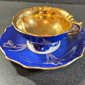 May include: A vintage teacup and saucer set. The teacup has a gold interior and a blue exterior with gold trim. The saucer is blue with gold floral accents and a gold rim. The set is likely porcelain.