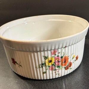 May include: Off-white ceramic baking dish with vertical ribbed sides and a floral design. The flowers are in shades of yellow, orange, pink, and red. The dish has a slightly angled rim.