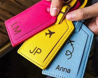 May include: Three colorful luggage tags are held in a person's hand. The tags are pink, yellow, and blue. The pink tag has the initials "LM", the yellow tag has "JP" and an airplane graphic, and the blue tag has the name "Anna" and an airplane graphic.