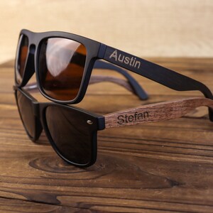 May include: Two pairs of sunglasses with black frames and dark lenses. The top pair has the name "Austin" engraved on the wooden arm. The bottom pair has the name "Stefan" engraved on the wooden arm.