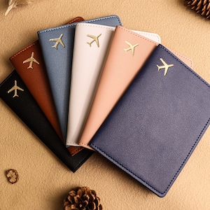 May include: Six passport holders in different colours, each with a gold airplane design. The colours are brown, blue, pink, white, and two shades of blue.