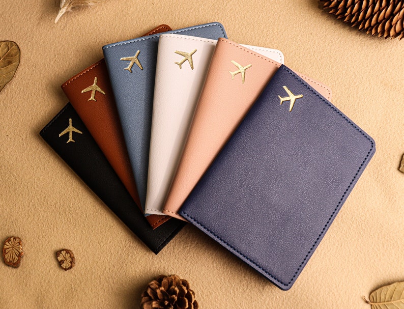 May include: Six passport holders in different colours, each with a gold airplane design. The colours are brown, blue, pink, white, and two shades of blue.