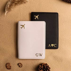 May include: Two passport holders, one white and one black, with gold airplane and heart designs. The white passport holder has the initials "GW" and the black passport holder has the initials "A&hearts;C".