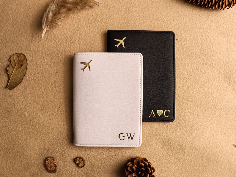 May include: Two passport holders, one white and one black, with gold airplane and heart designs. The white passport holder has the initials "GW" and the black passport holder has the initials "A&hearts;C".