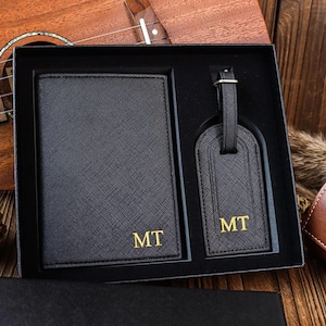 K&ouml;nnte beinhalten: Ein schwarzes Leder-Reisepass-Etui und ein Gep&auml;ckanh&auml;nger-Set mit goldenen Buchstaben. Das Set ist mit den Initialen "MT" monogrammiert.
