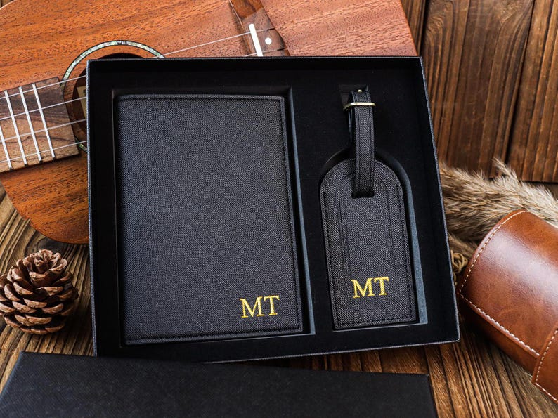 K&ouml;nnte beinhalten: Ein schwarzes Leder-Reisepass-Etui und ein Gep&auml;ckanh&auml;nger-Set mit goldenen Buchstaben. Das Set ist mit den Initialen "MT" monogrammiert.