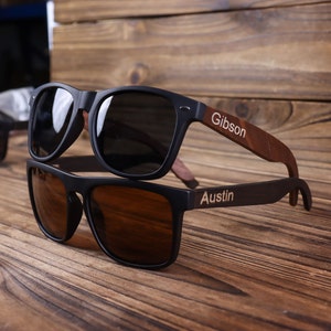 May include: Two pairs of black sunglasses with wooden arms. The top pair has the name "Gibson" engraved on the arm. The bottom pair has the name "Austin" engraved on the arm.