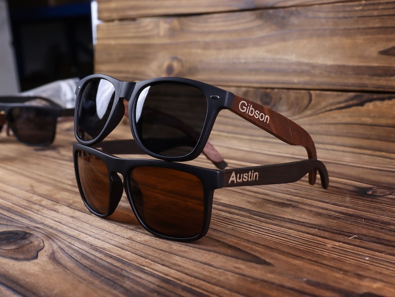 May include: Two pairs of black sunglasses with wooden arms. The top pair has the name "Gibson" engraved on the arm. The bottom pair has the name "Austin" engraved on the arm.