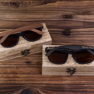 May include: Two pairs of wooden sunglasses in their individual boxes. The sunglasses have black frames and brown lenses. The boxes are made of wood and have a light brown finish. The box on the left has the name "Austin" engraved on it. The box on the right has the words "Best Man" engraved on it.