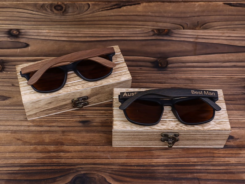 May include: Two pairs of wooden sunglasses in their individual boxes. The sunglasses have black frames and brown lenses. The boxes are made of wood and have a light brown finish. The box on the left has the name "Austin" engraved on it. The box on the right has the words "Best Man" engraved on it.