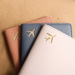 May include: Three passport holders in different colours: pink, blue, and light pink. Each holder has a gold airplane design.