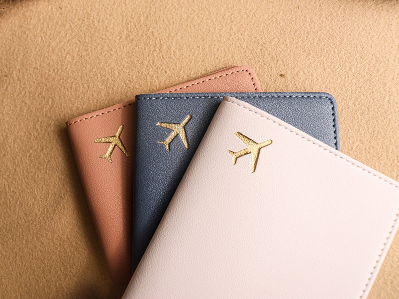 May include: Three passport holders in different colours: pink, blue, and light pink. Each holder has a gold airplane design.
