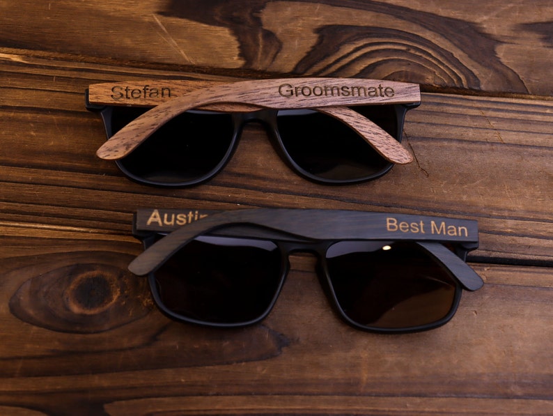 May include: Two pairs of sunglasses with wooden frames. The top pair has a brown frame with the word "Groomsmate" engraved on the right side and the word "Stefan" engraved on the left side. The bottom pair has a black frame with the word "Best Man" engraved on the right side and the word "Austin" engraved on the left side.