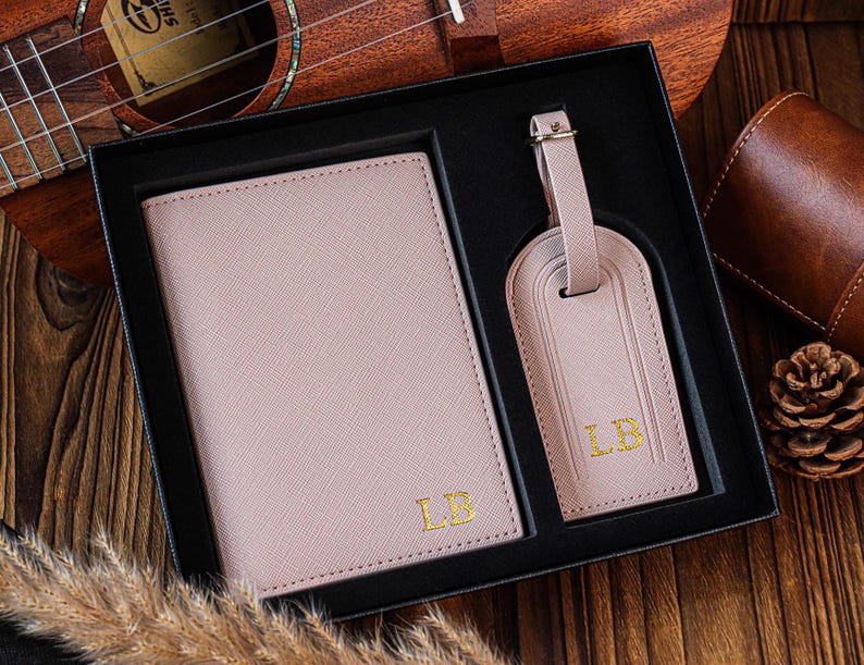 K&ouml;nnte beinhalten: Ein Set aus einem rosafarbenen Leder-Reisepass-Etui und einem Gep&auml;ckanh&auml;nger mit goldenen Initialen "LB" auf jedem Teil. Das Set wird in einer schwarzen Box pr&auml;sentiert.