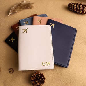 May include: Five passport holders in different colours, including navy blue, light pink, brown, light blue, and peach. Each holder has a gold airplane design and the pink holder has the initials "GW" embossed on the front.