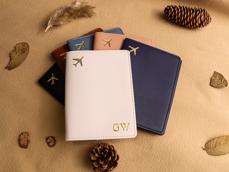 May include: Five passport holders in different colours, including navy blue, light pink, brown, light blue, and peach. Each holder has a gold airplane design and the pink holder has the initials "GW" embossed on the front.