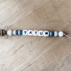 May include: A baby pacifier clip with the name "TATUM" spelled out in white square beads with black letters. The clip has a metal clasp and is strung with gray, blue, and white beads.