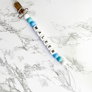 May include: A baby pacifier clip with the name "WALKER" spelled out in white letter beads. The clip features a gold-colored metal clasp and a series of blue, teal, and white beads. A clear loop is attached at the end.