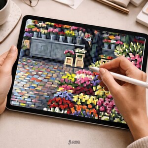May include: A tablet displaying a pixelated digital illustration of a flower shop with a cobblestone street, colorful floral arrangements, and a storefront. A white stylus is held against the screen, which also features a small logo at the bottom.