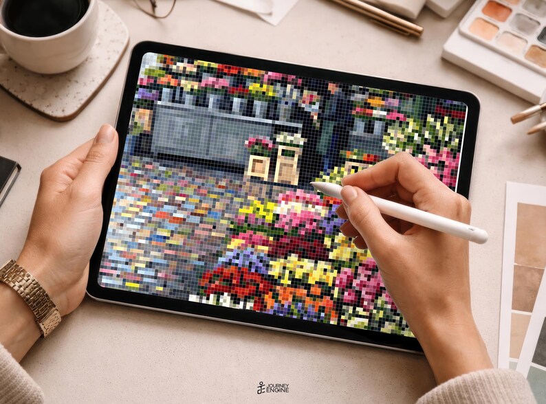 May include: A tablet displaying a pixelated digital illustration of a flower shop with a cobblestone street, colorful floral arrangements, and a storefront. A white stylus is held against the screen, which also features a small logo at the bottom.